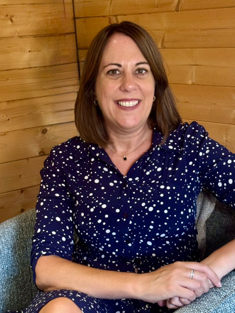 Me in my counselling room. Blue garden shed with a deck, chairs, and potted plants in a lush green setting.